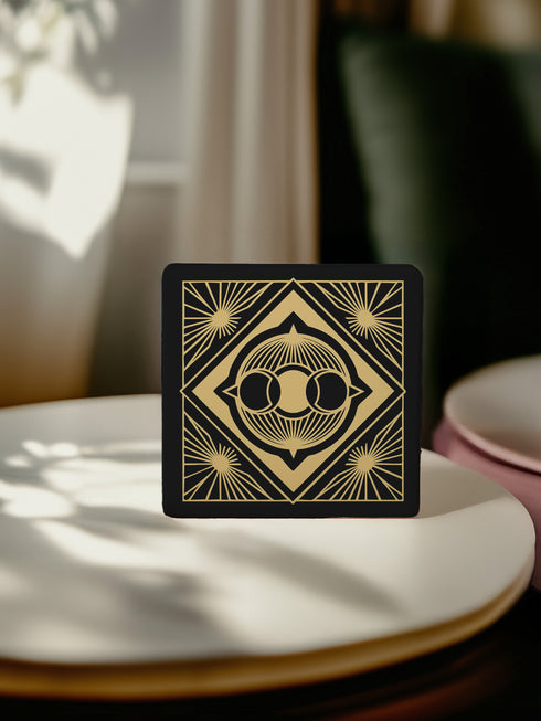 A black and gold ceramic coaster featuring a triple moon design framed by art deco geometric details and celestial sunburst accents.